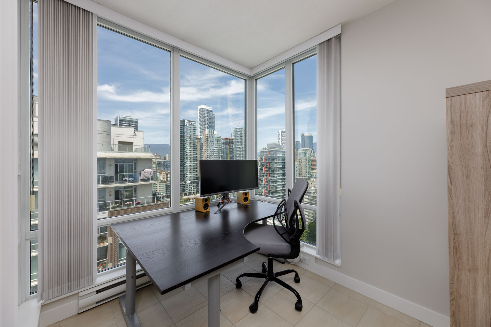 Home office with a computer monitor, speakers, and chair positioned by large windows overlooking a cityscape with high-rise buildings.