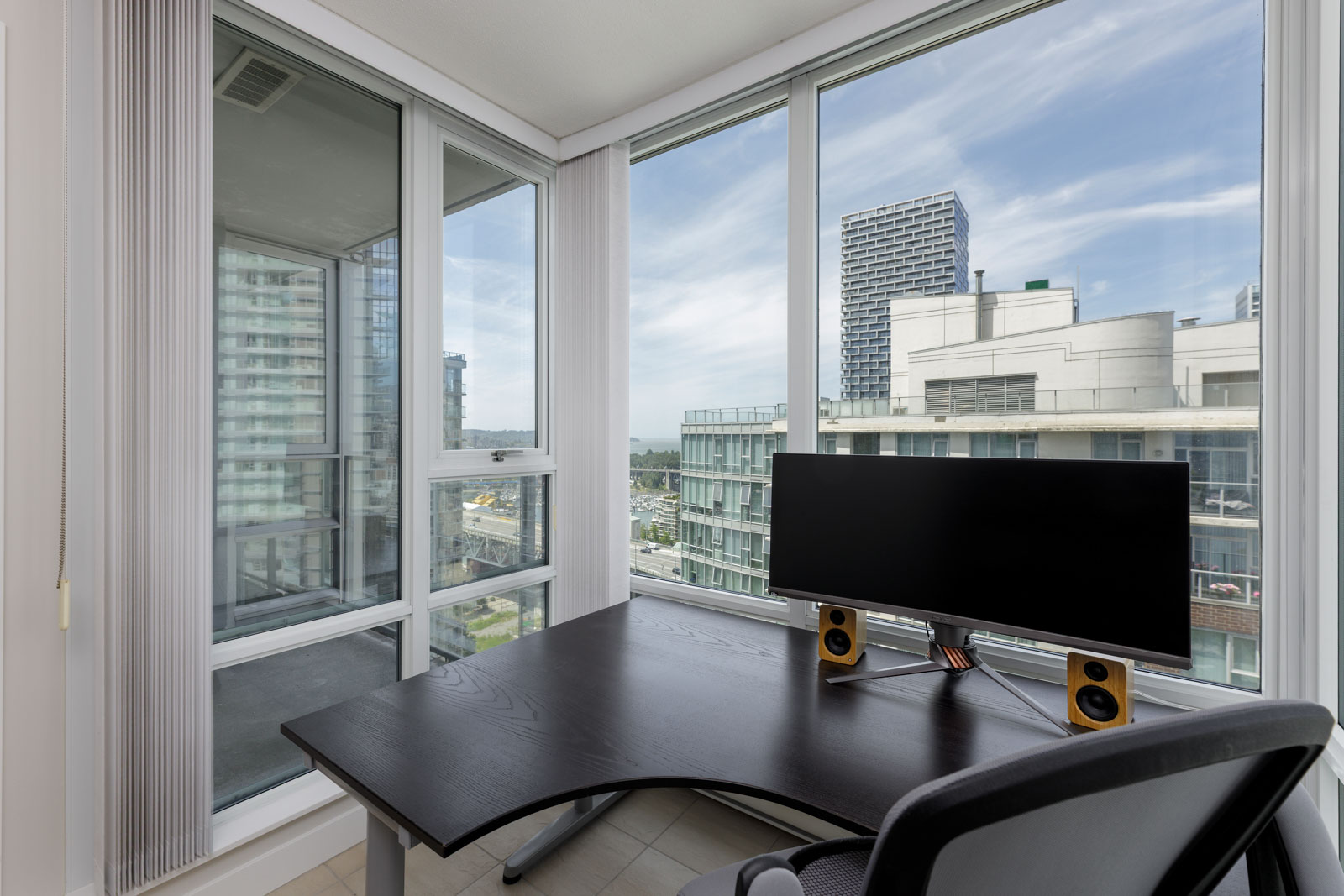 Modern corner office with large windows, a black desk, computer monitor, speakers, and cityscape view with high-rise buildings in the background.