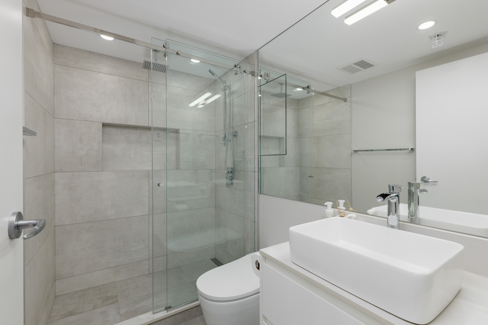 Modern bathroom with large glass shower enclosure, wall-mounted toilet, rectangular sink, and neutral-colored tiles.