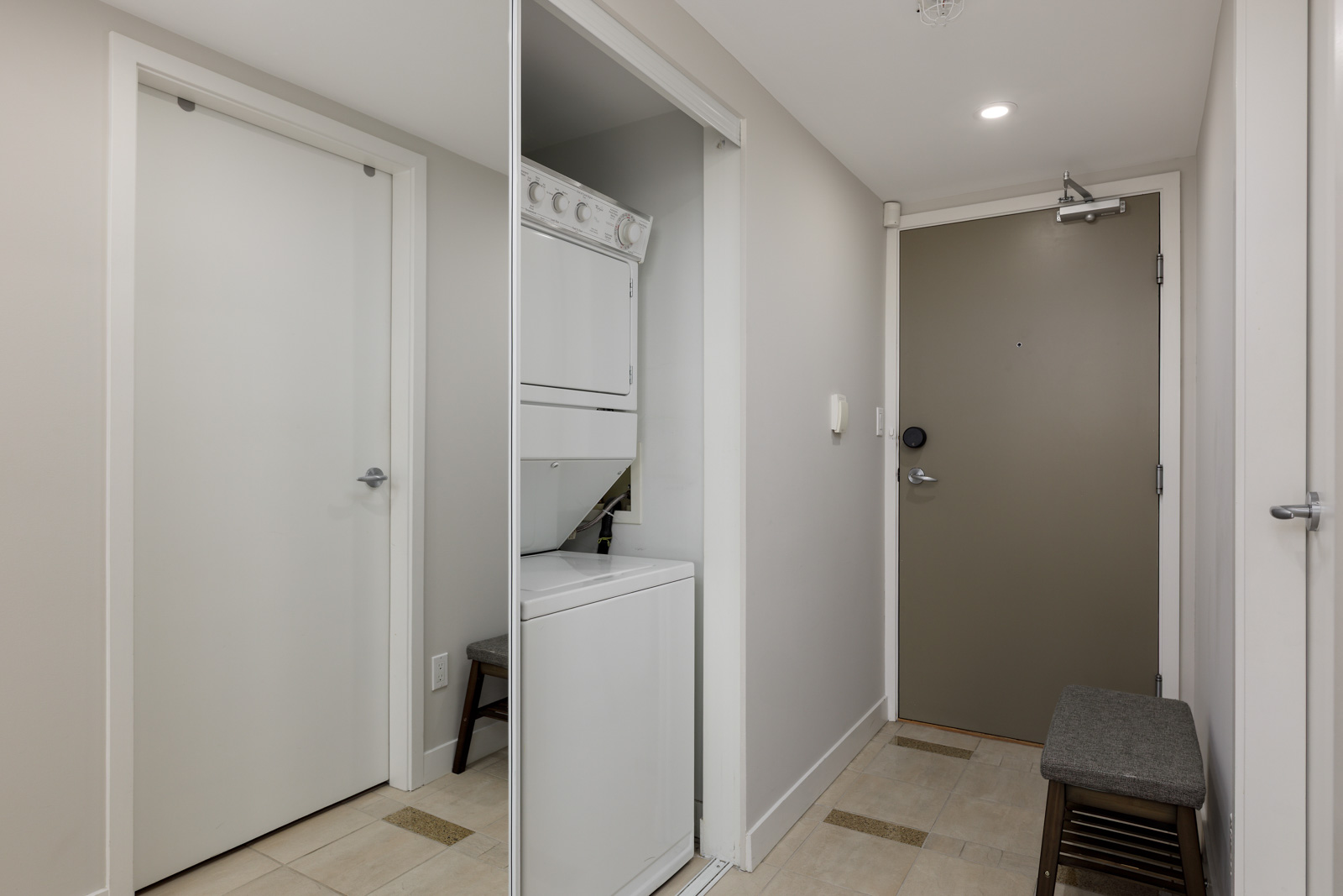 A hallway with a closed door, a front door, a small bench, and a stacked washer and dryer unit in an open closet.