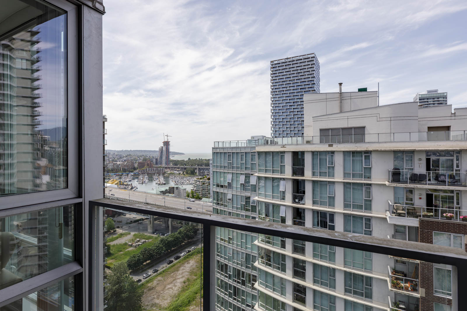 View from a balcony overlooking modern apartment buildings, a road, and a distant waterfront under a partly cloudy sky.