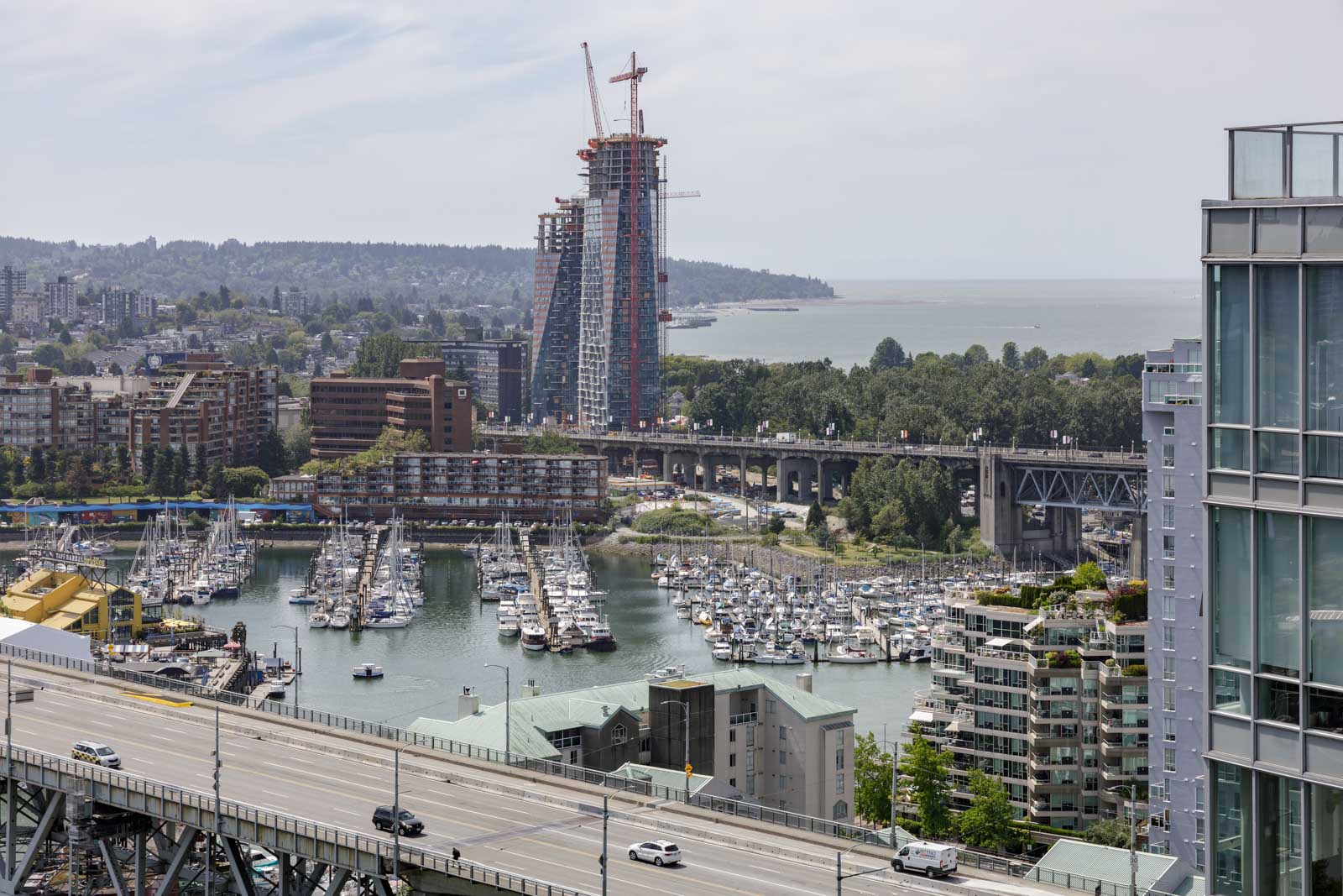 A marina filled with boats beside a highway overpass, with high-rise buildings and a tall construction crane in the background near a body of water.