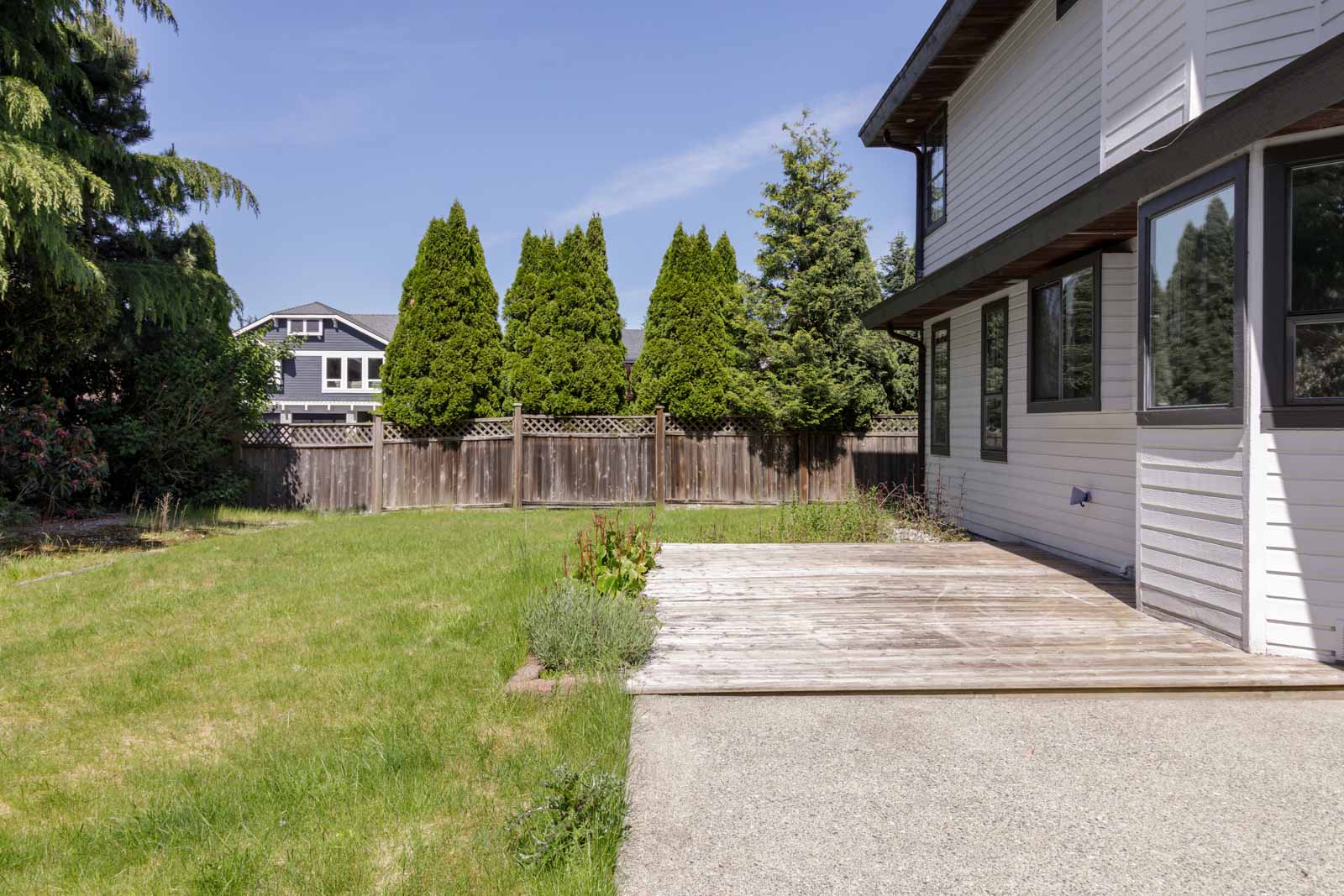 A backyard with a grassy lawn, wooden patio, and partial view of a white house; wooden fence and tall trees line the perimeter.