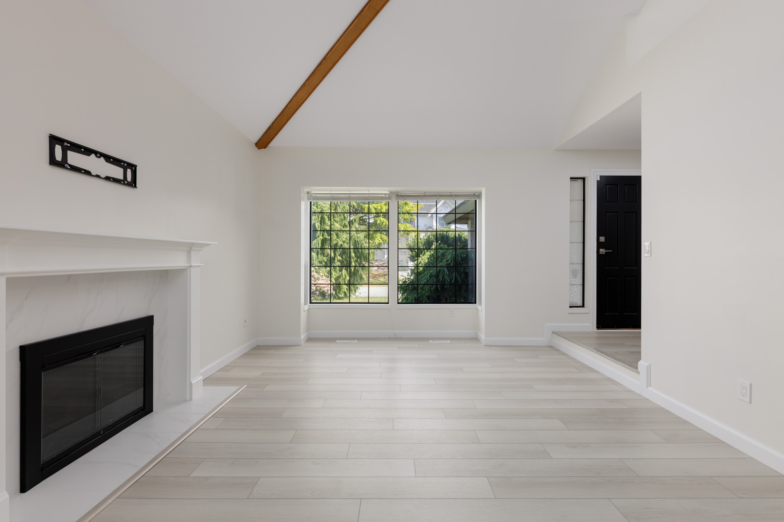 Bright, empty living room with light wood flooring, white walls, a fireplace, large windows, and a black front door.