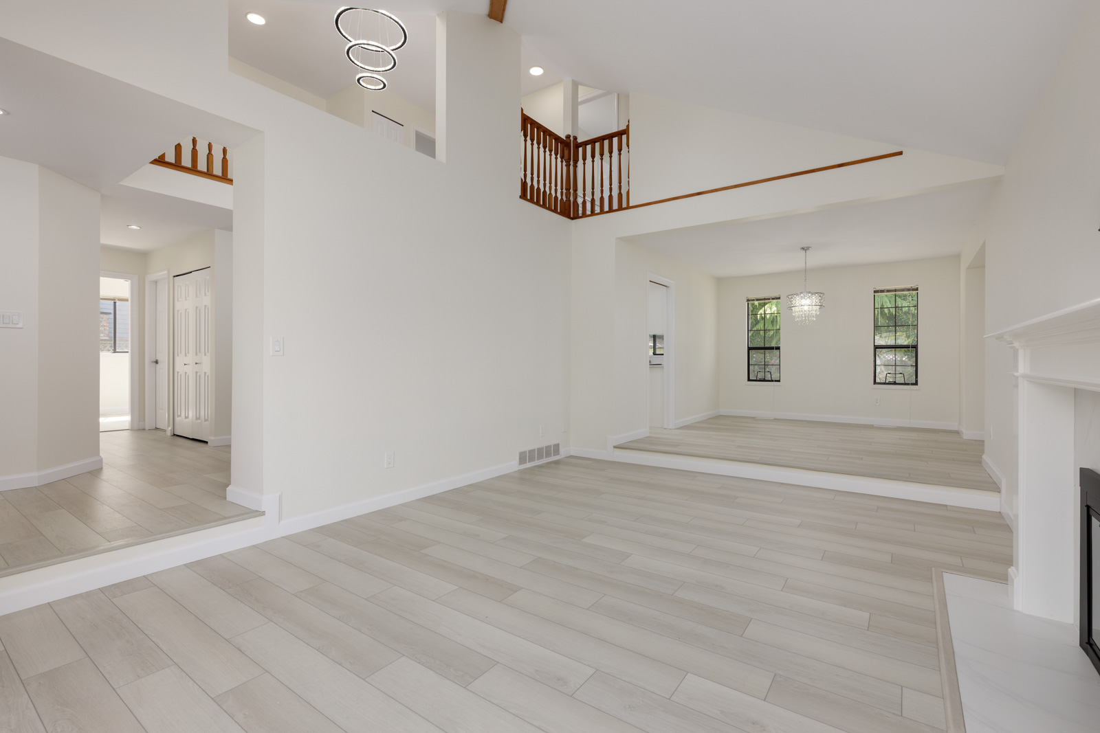 Spacious modern living room with light wood flooring, white walls, high ceilings, a fireplace, and large windows; staircase and chandelier visible in adjacent areas.