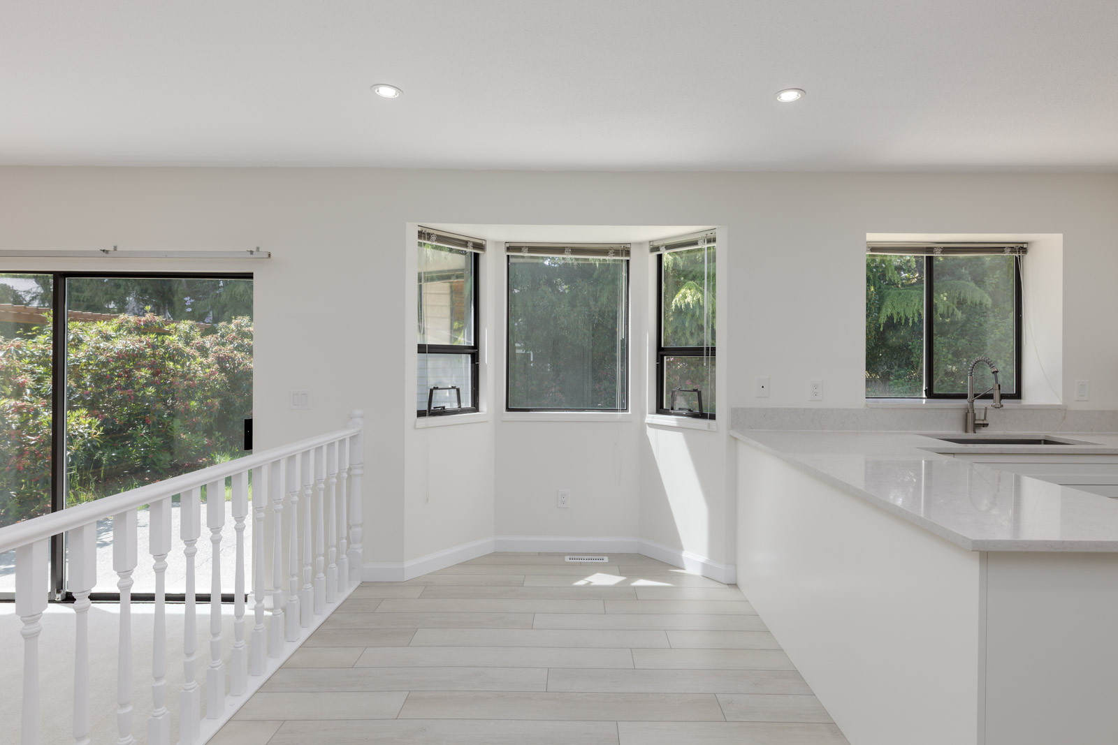 Bright kitchen area with white countertops, light tile flooring, multiple windows, and a sliding glass door leading to a patio with greenery outside.