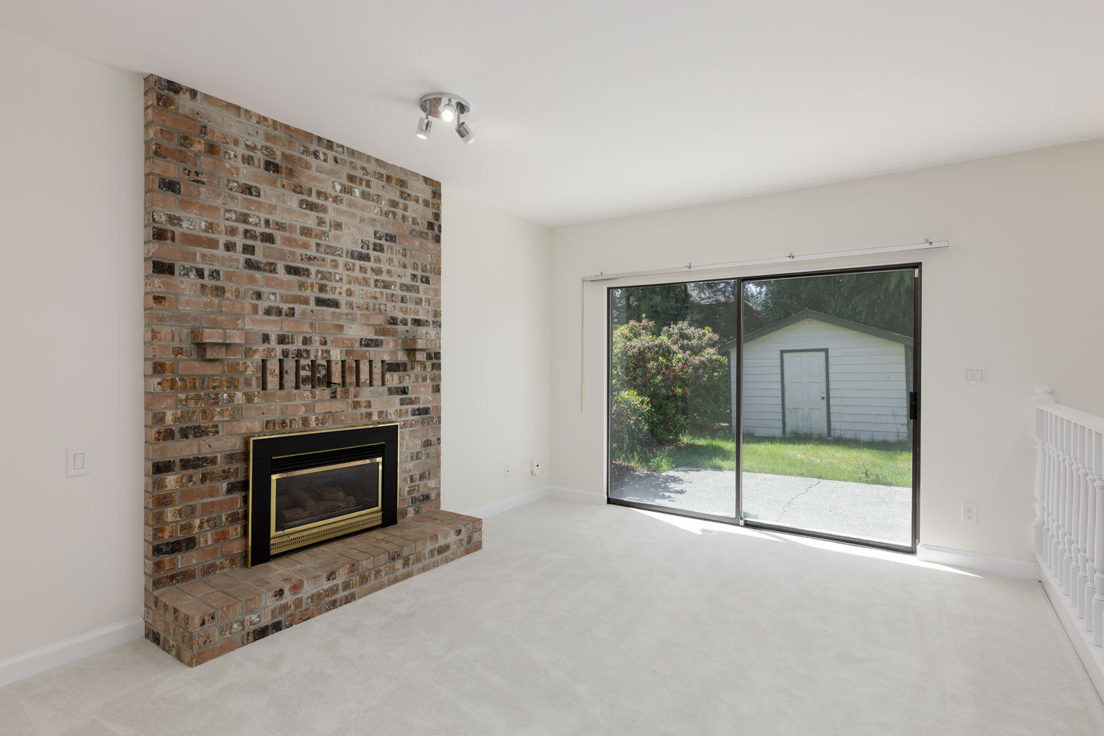 A living room with a brick fireplace, beige carpet, and large sliding glass doors leading to a backyard with a shed.