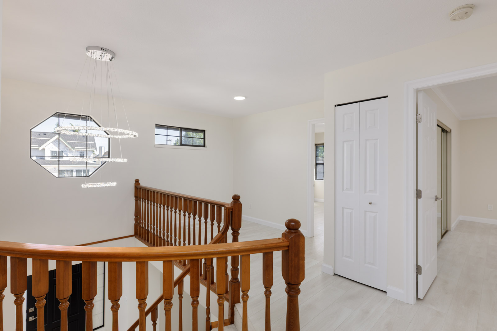 A bright upstairs hallway with wooden railing, modern chandelier, light-colored walls, and doors leading to adjacent rooms.