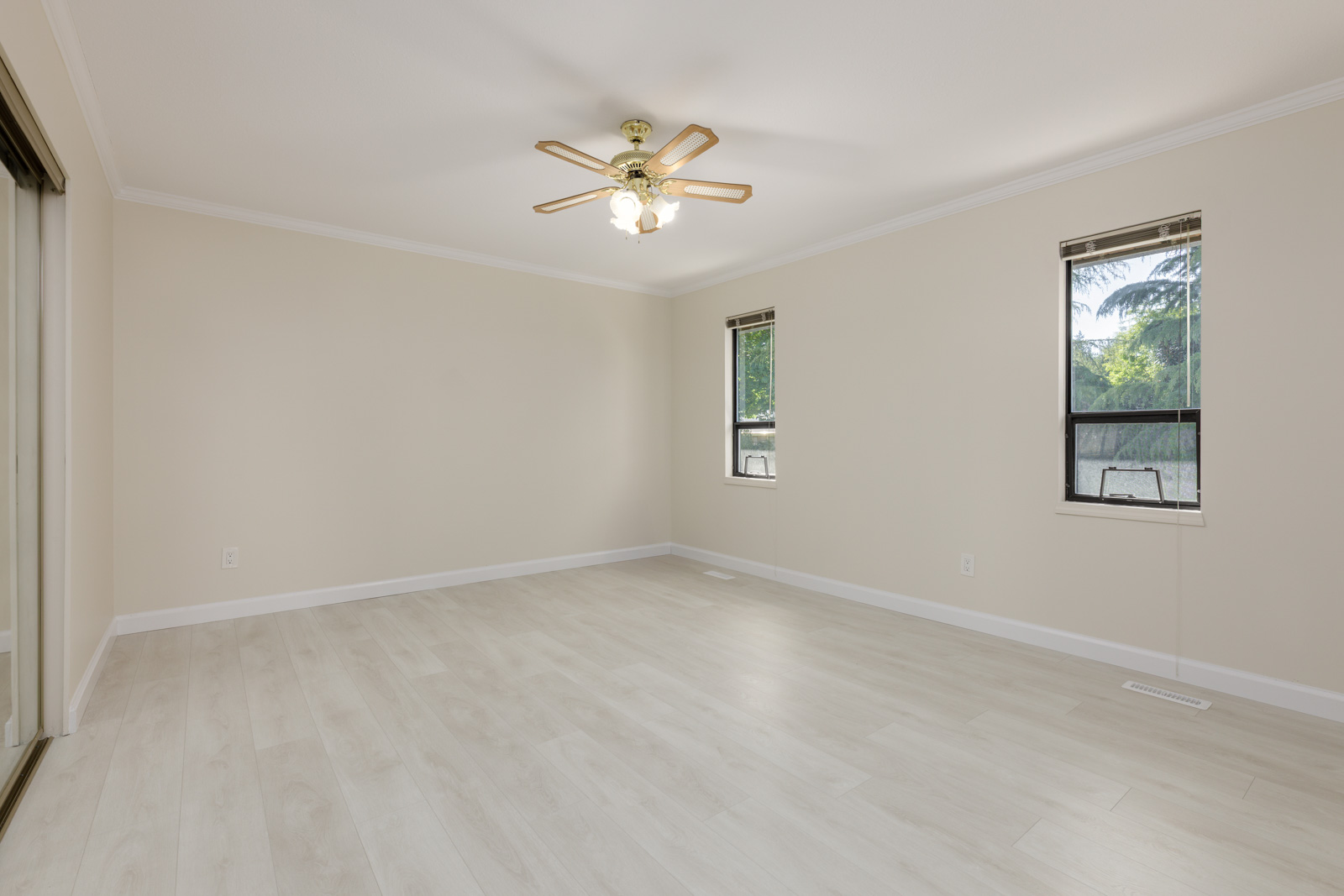 Empty room with light-colored wood flooring, beige walls, two windows, a ceiling fan with lights, and mirrored closet doors on the left.