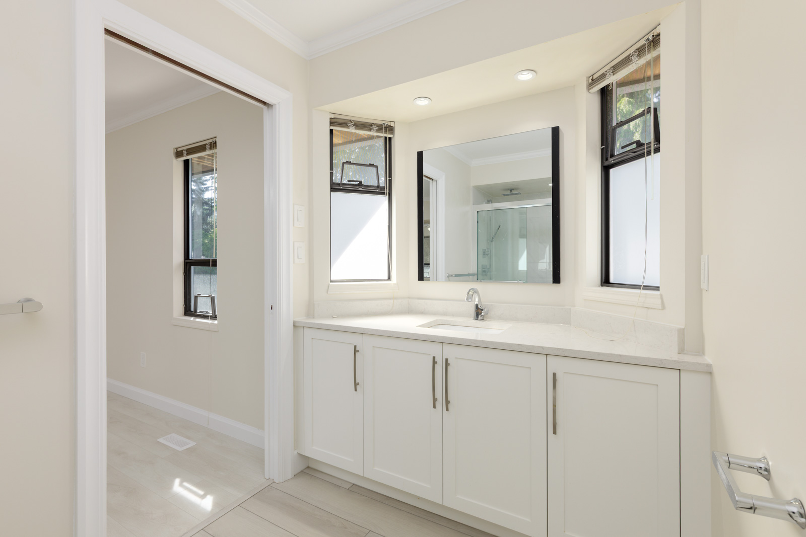 Modern bathroom with white cabinets, a single sink, large mirror, and two windows; view into adjacent room with similar flooring and wall color.