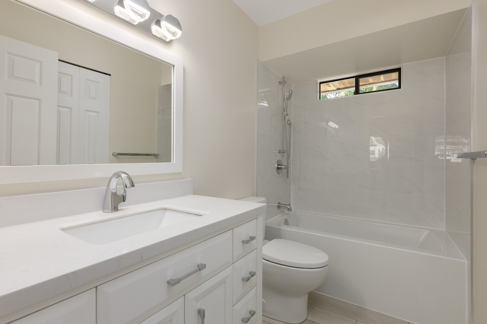 Modern bathroom with white cabinets, a sink, a toilet, and a bathtub-shower combo; rectangular window above tub and bright vanity lighting.
