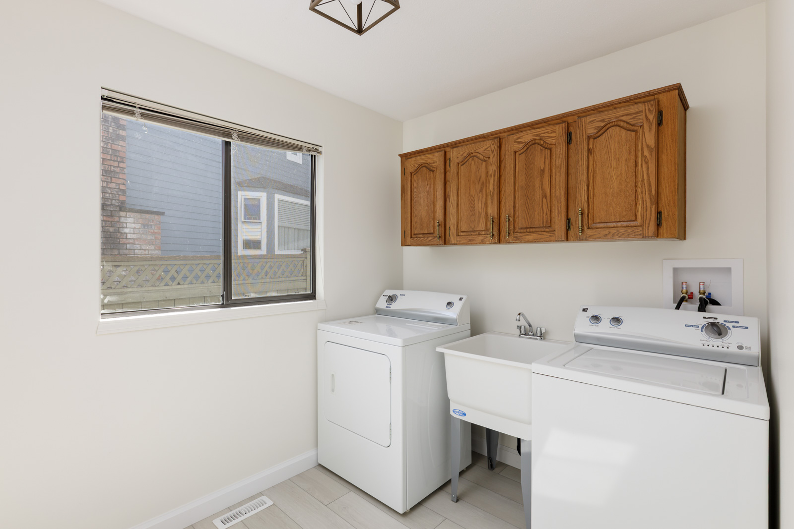 A laundry room with a washer, dryer, utility sink, wooden wall cabinets, and a window with blinds.