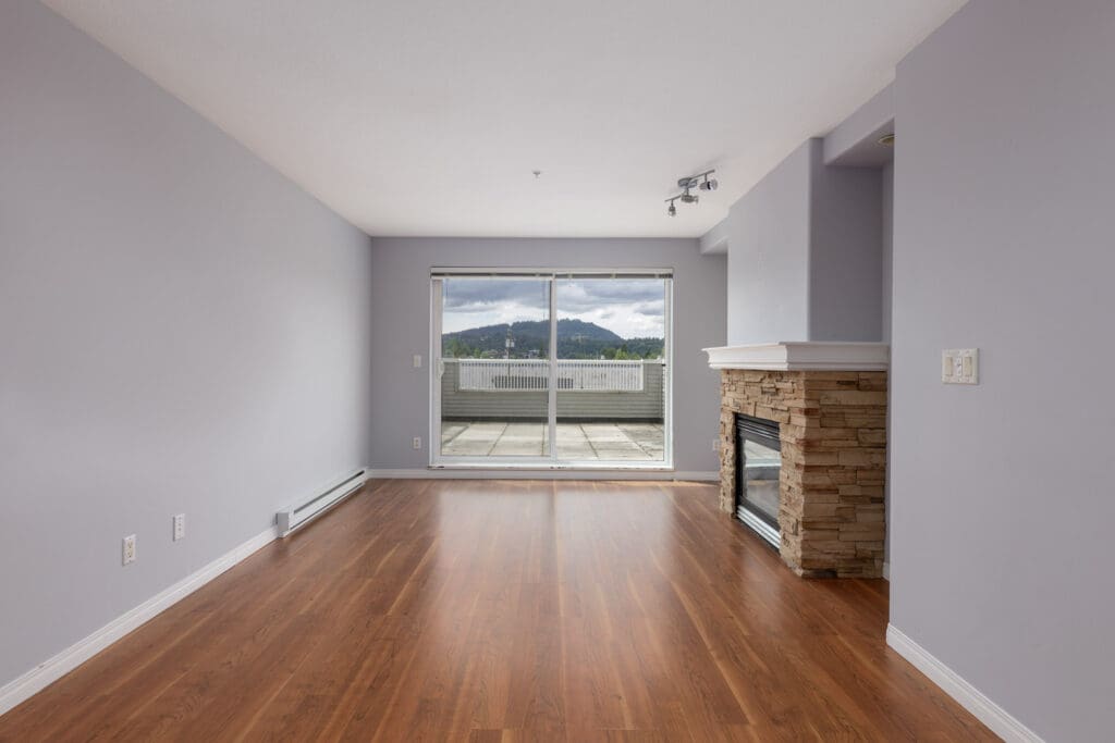 Empty living room with light purple walls, wooden flooring, a stone fireplace, and large sliding glass doors leading to a balcony with a mountain view.