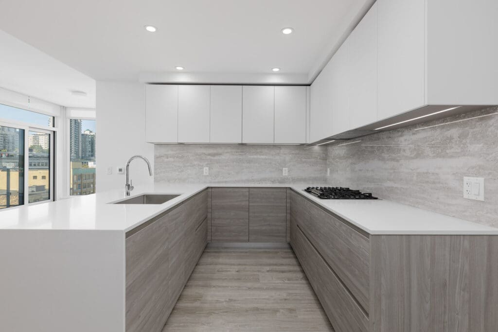 Modern, minimalist kitchen with white countertops, light wood cabinets, built-in gas stove, sink, and large windows providing city views.