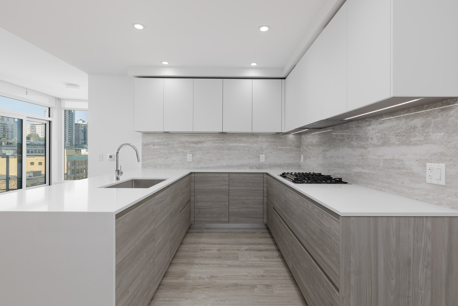 Modern, minimalist kitchen with white countertops, light wood cabinets, built-in gas stove, sink, and large windows providing city views.