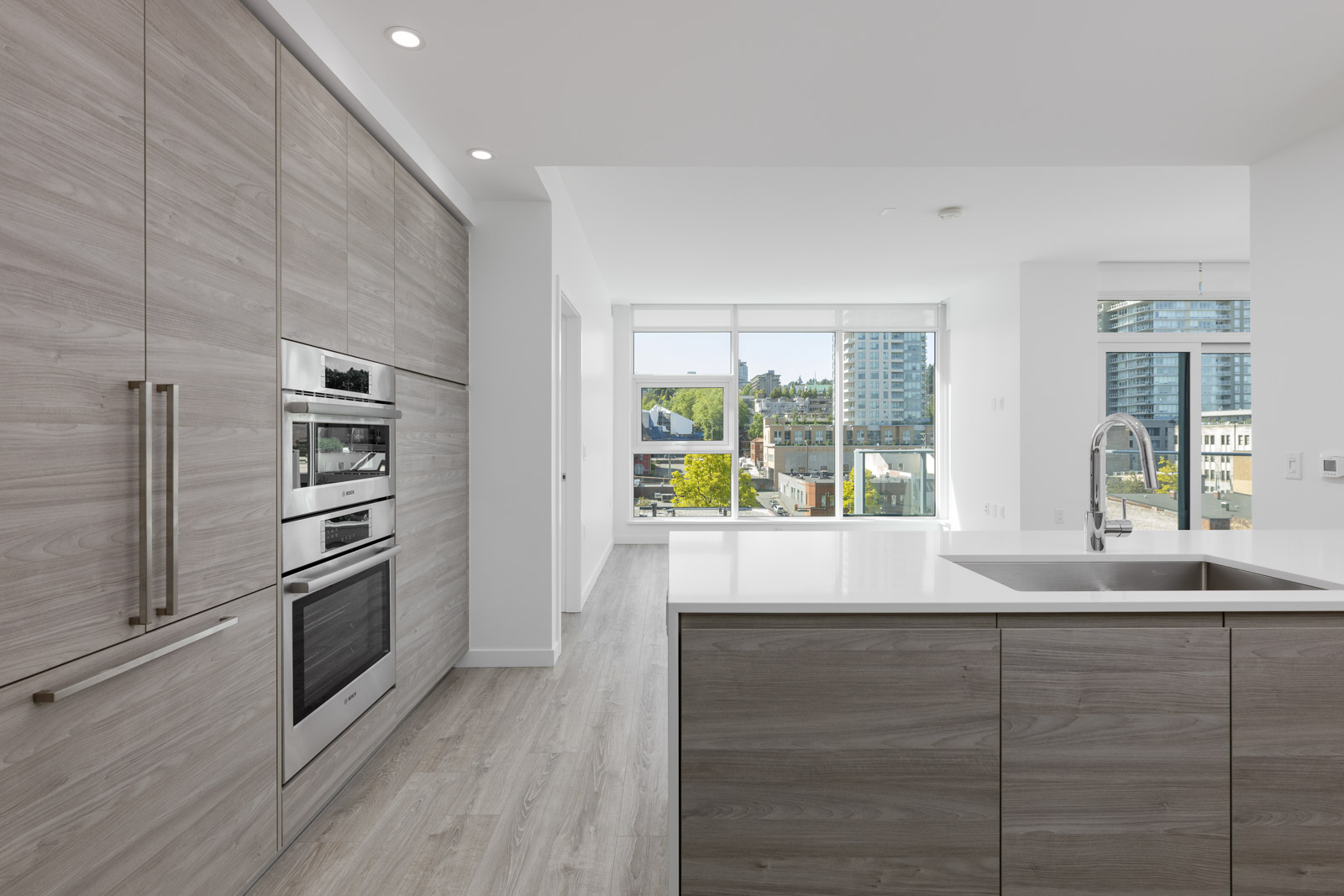 Modern kitchen with light wood cabinets, built-in stainless steel appliances, island with sink, and large windows overlooking a cityscape.