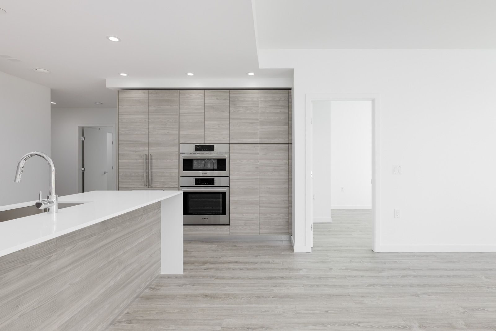 Modern kitchen with light wood cabinets, built-in stainless steel appliances, a white countertop, and adjacent open doorway leading to a bright, unfurnished room.