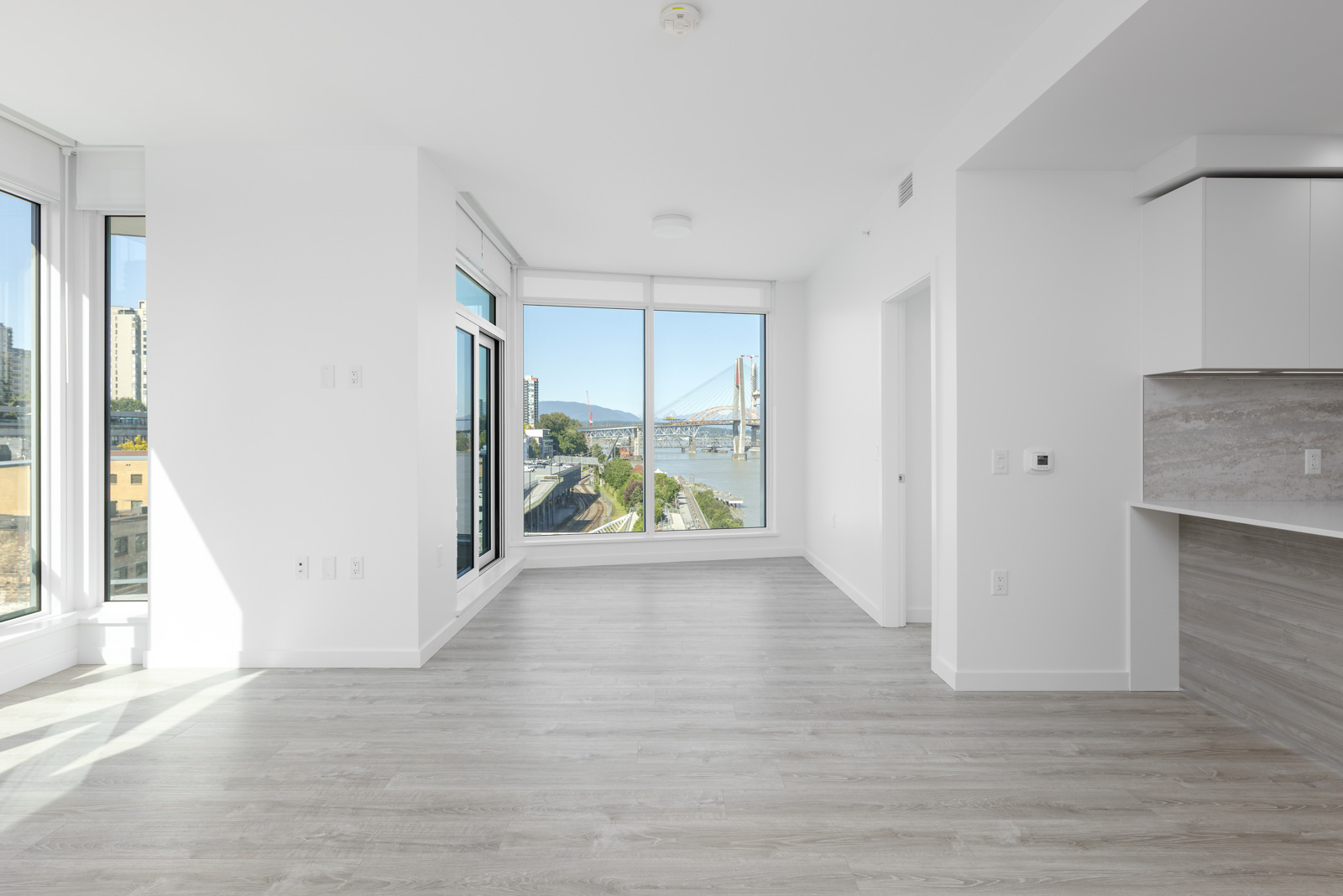 Empty modern apartment with large windows, light gray flooring, white walls, and a view of a river and bridge outside.
