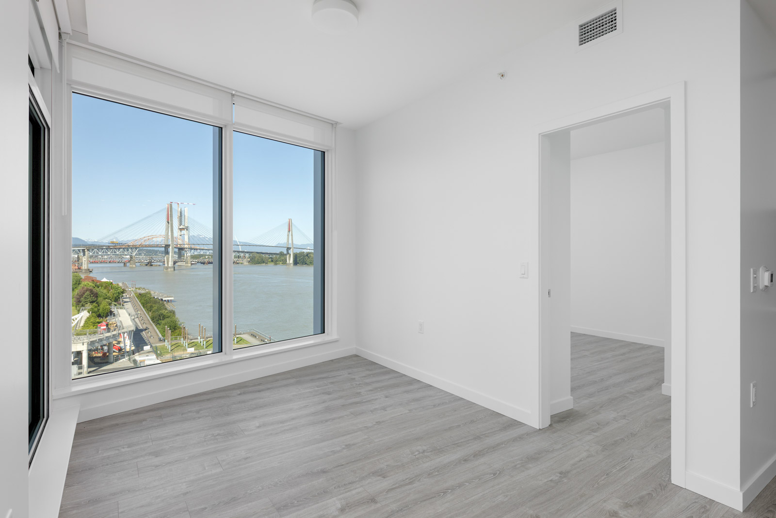 Unfurnished room with large windows overlooking a river and bridge, white walls, light gray wood flooring, and an open doorway leading to another room.