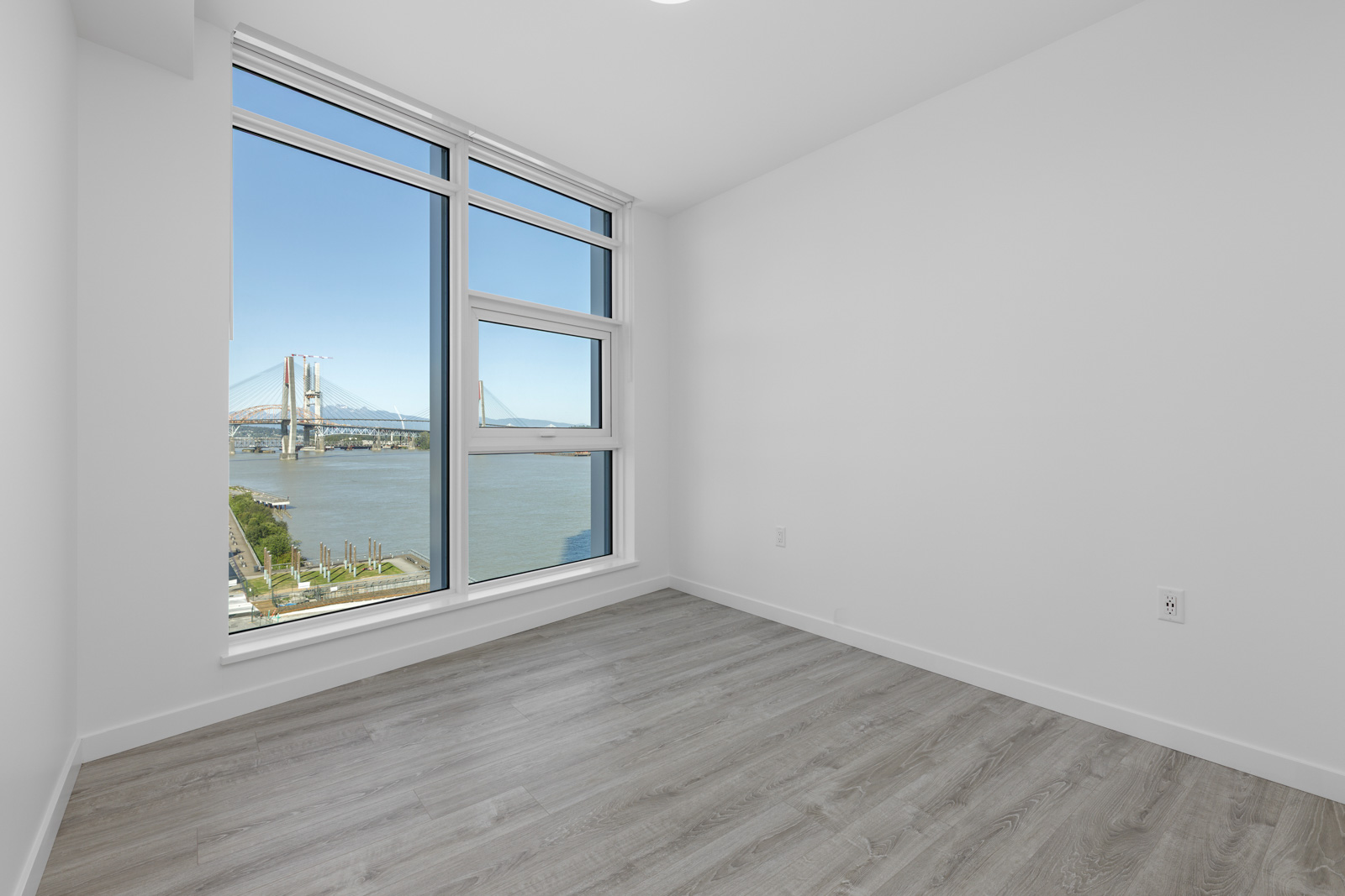 Empty room with light gray wood flooring and white walls, featuring a large window with a view of a river and a suspension bridge outside.