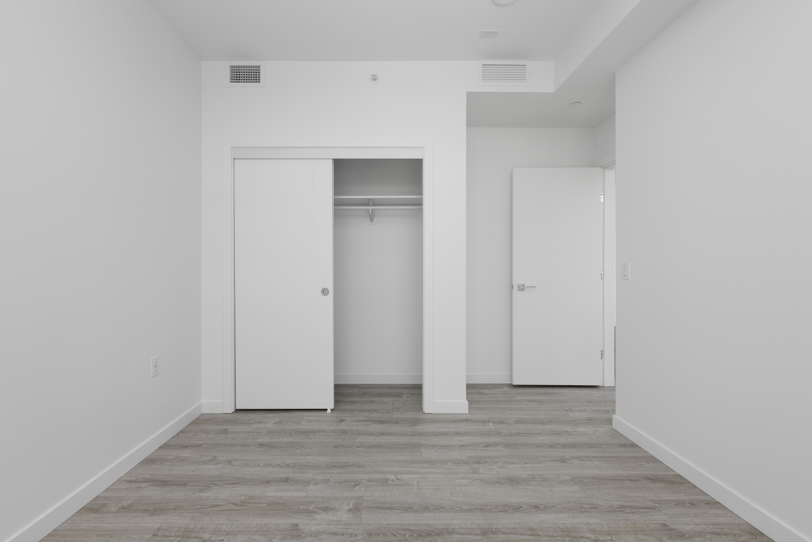 Empty room with light gray wood flooring, white walls, an open closet with sliding doors, and an open door leading to another area.