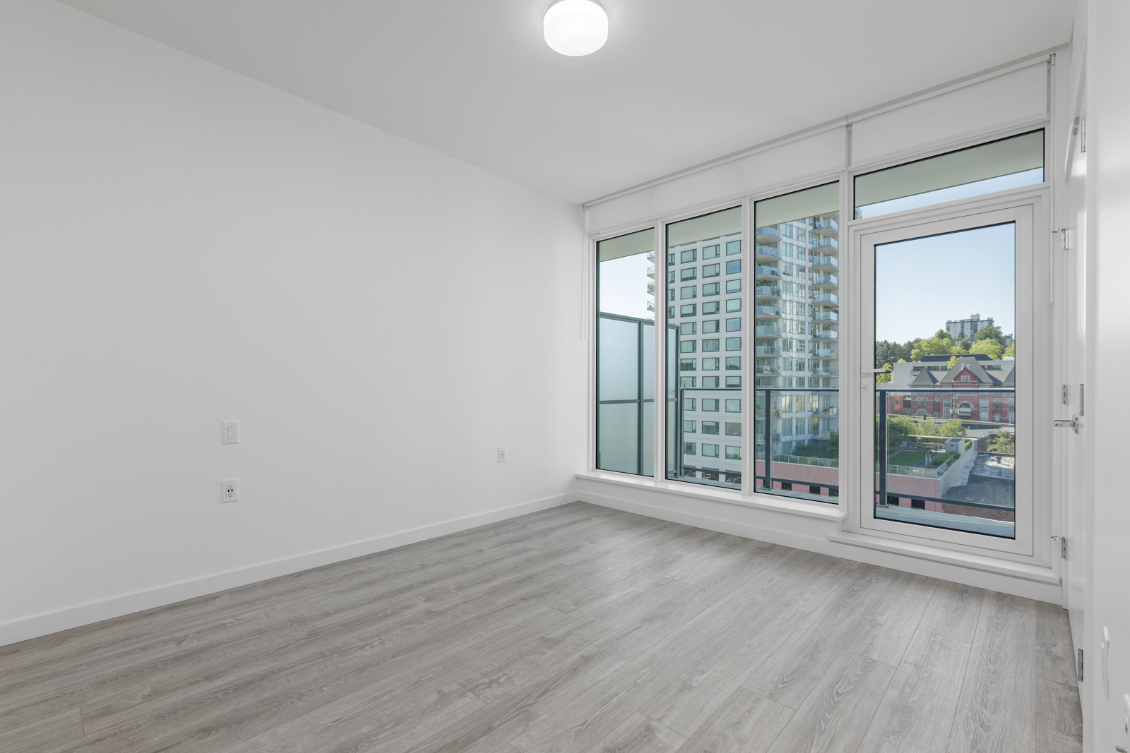 Empty modern room with light wood flooring, white walls, and large windows offering a view of city buildings and a balcony.