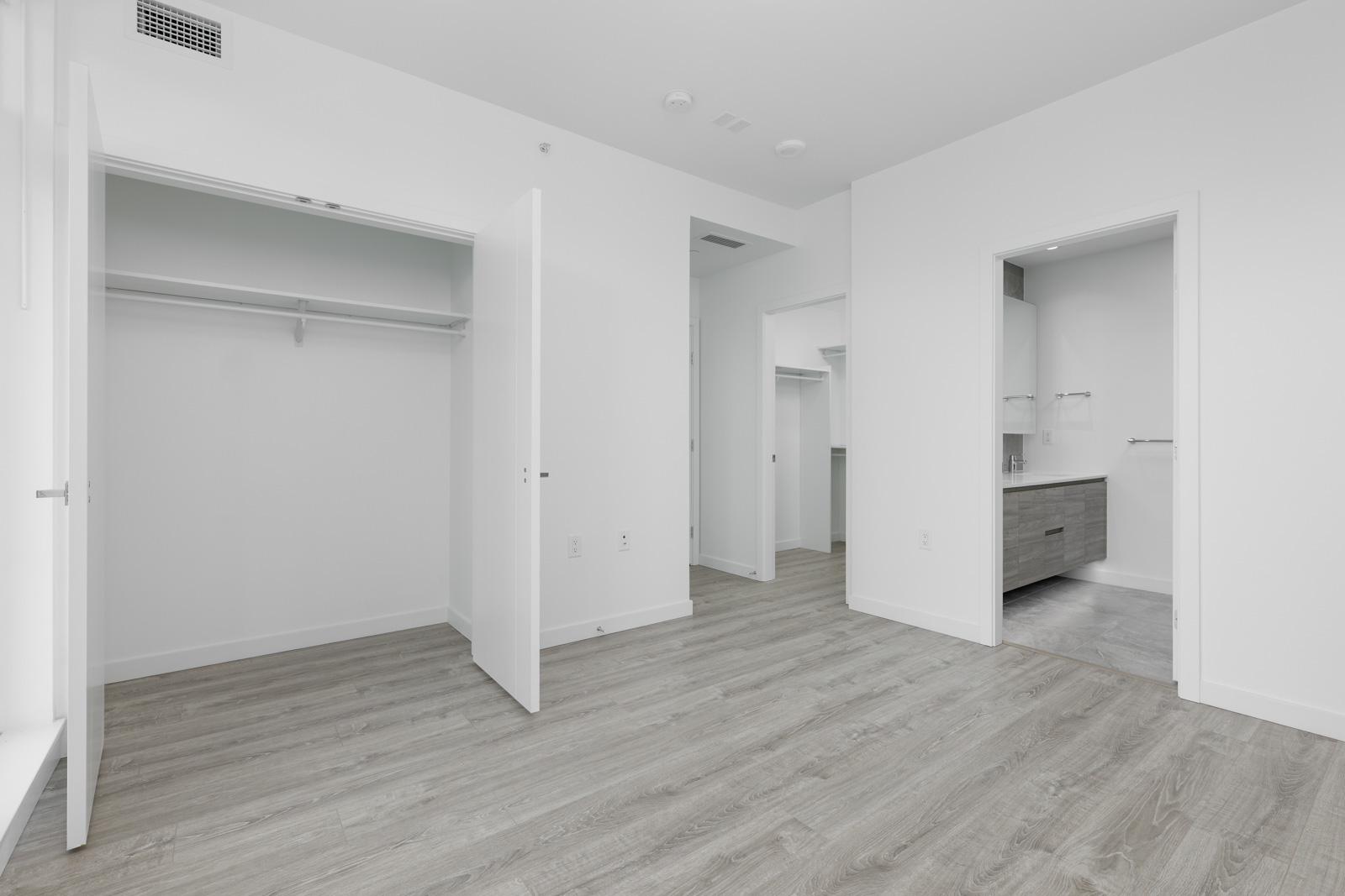 Empty modern bedroom with open closet, light wood flooring, white walls, and a view into a bathroom with a vanity.
