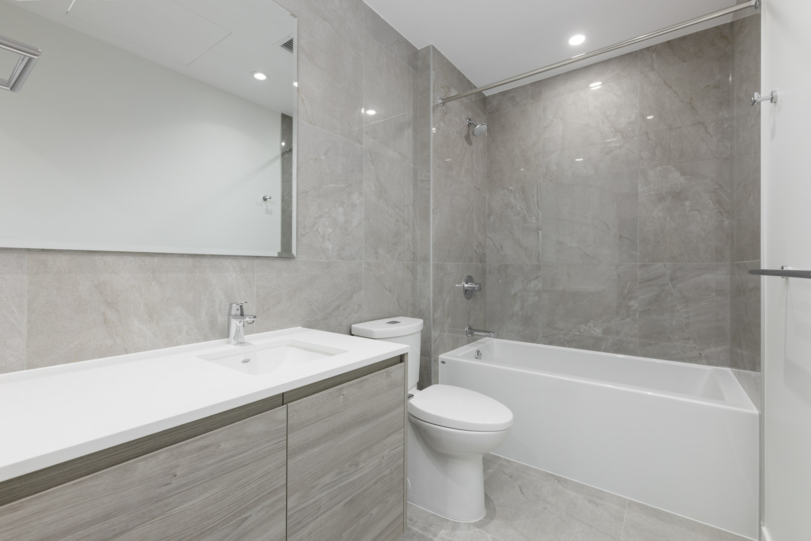 Modern bathroom with a large mirror, single sink vanity, toilet, and bathtub with shower, featuring light gray marble-style tiles on the walls and floor.