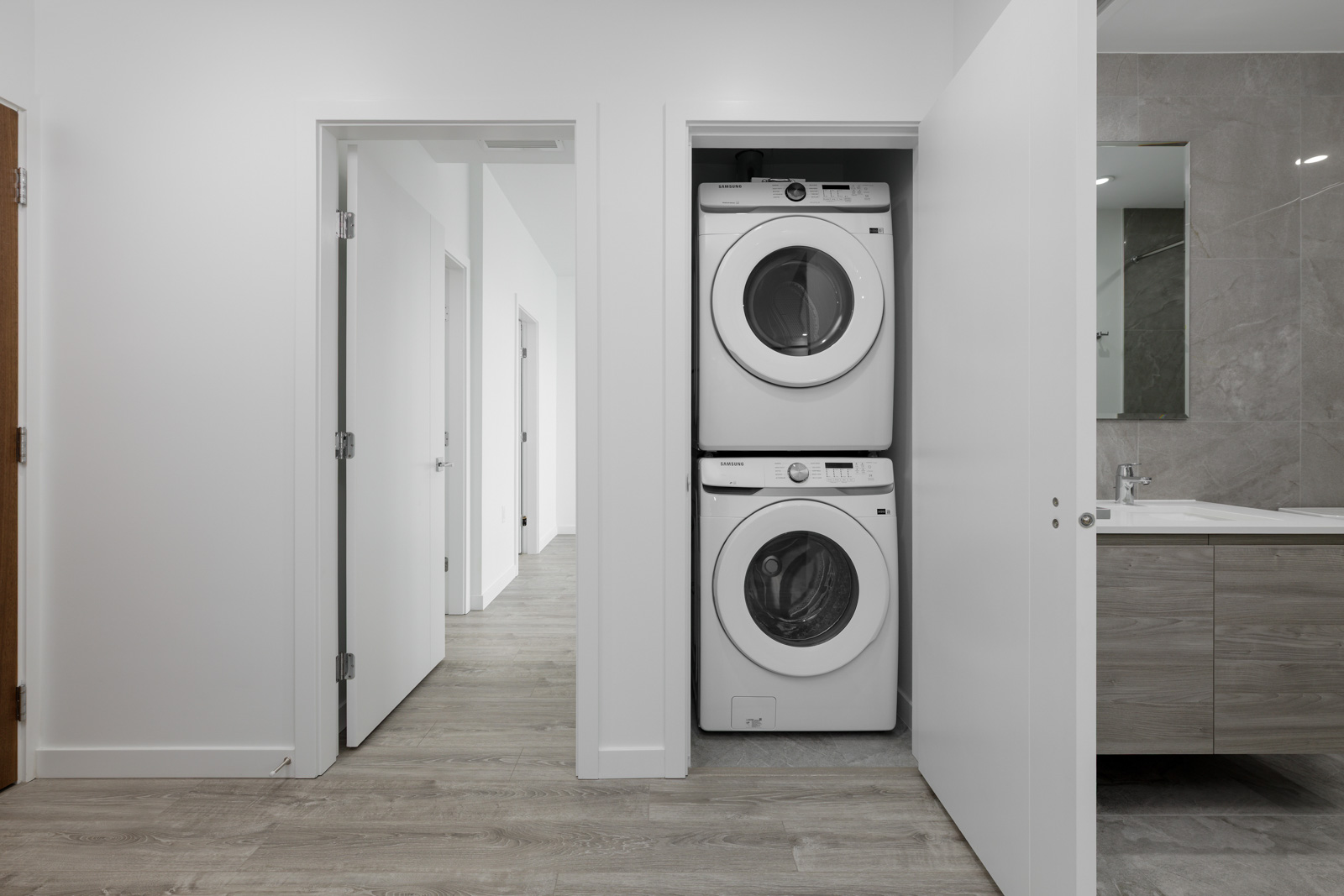 A stacked washer and dryer are placed in a closet next to a bathroom with a modern sink and mirror, in a hallway with light wood flooring and white walls.