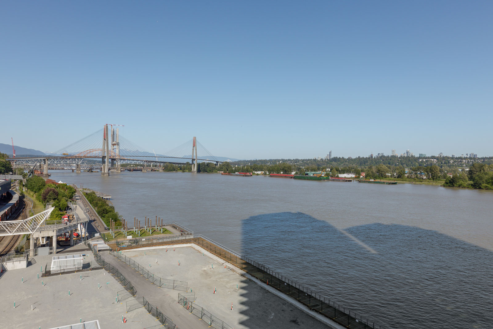 Wide view of a river with a large bridge spanning across, bordered by greenery and urban infrastructure under a clear blue sky.