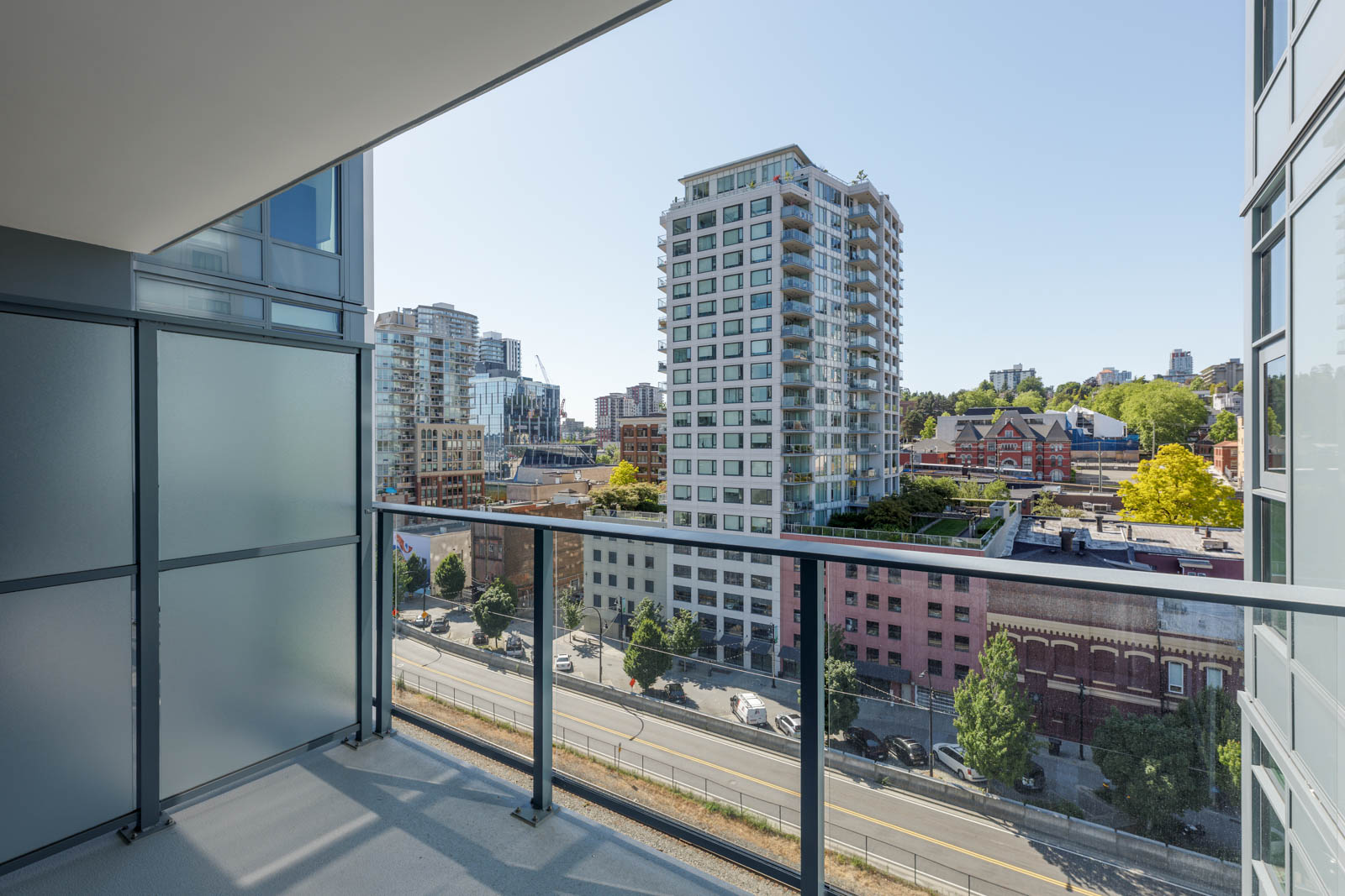 View from a modern balcony overlooking a city street, with mid-rise buildings, trees, and clear skies in the background.