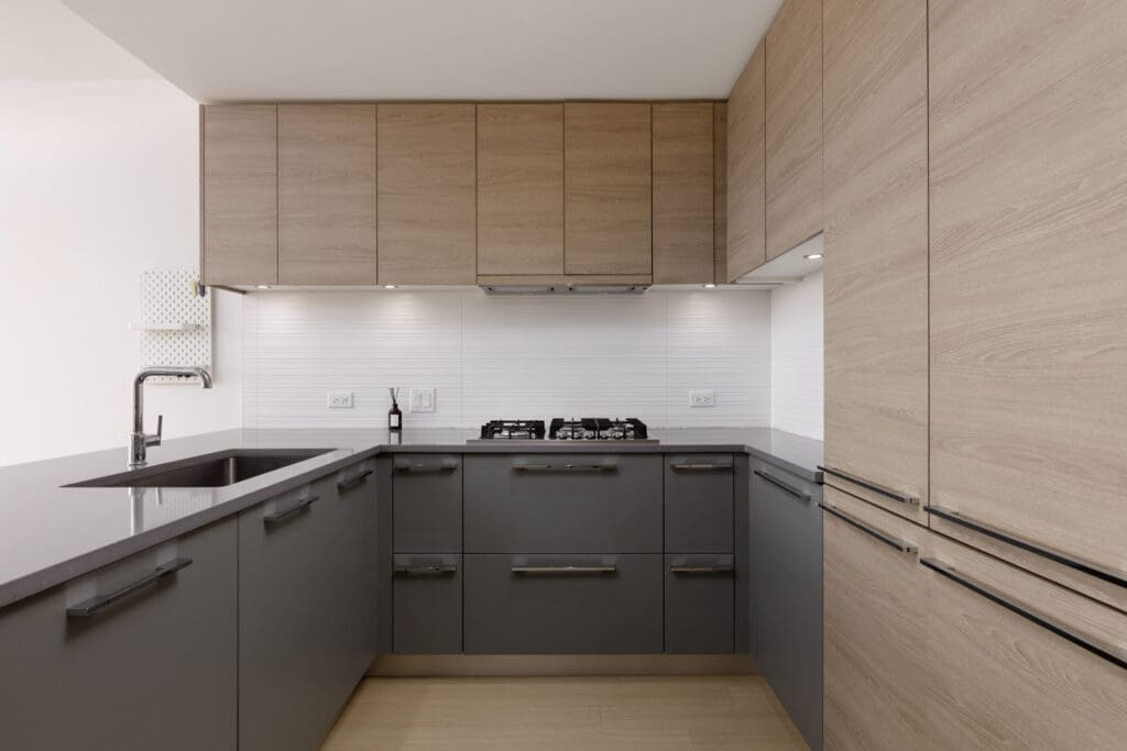 Modern kitchen with light wood upper cabinets, dark gray lower cabinets, a built-in gas stove, under-cabinet lighting, and a sink with a sleek faucet on a gray countertop.