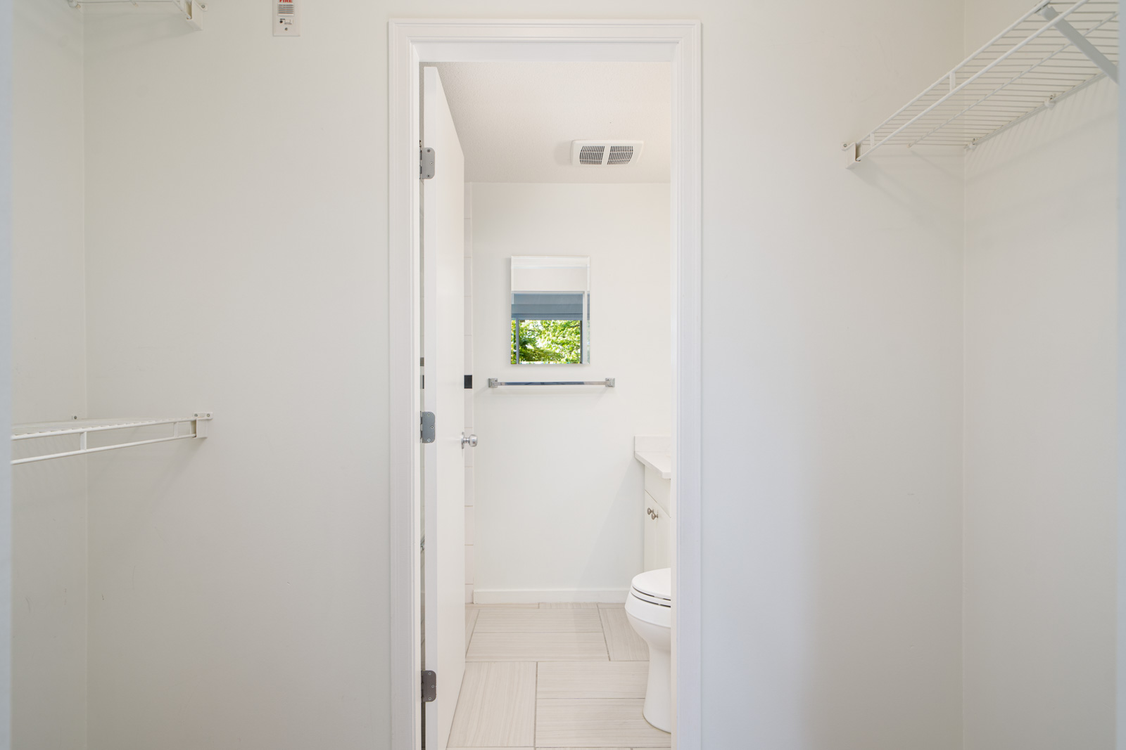View of a small, white bathroom with a toilet, countertop, wall-mounted mirror, and a window with greenery visible outside. A closet with wire shelving is in the foreground.