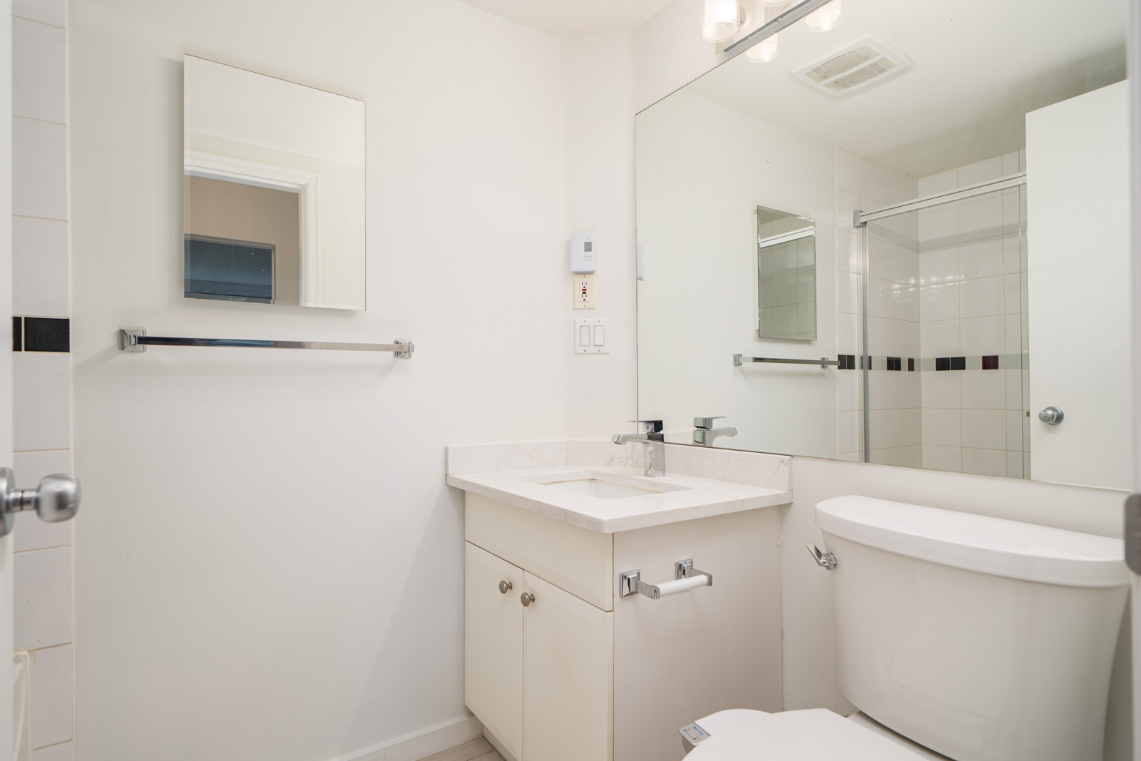 Modern bathroom with white walls, a vanity with sink, wall mirror, toilet, towel bar, and shower with glass door and tiled wall.