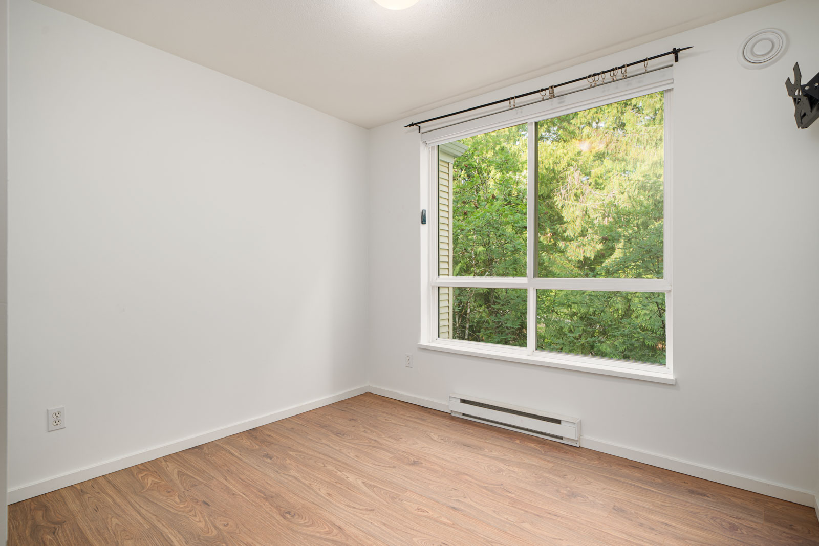 Unfurnished room with white walls, wood floor, large window with a curtain rod, baseboard heater, and view of green trees outside.