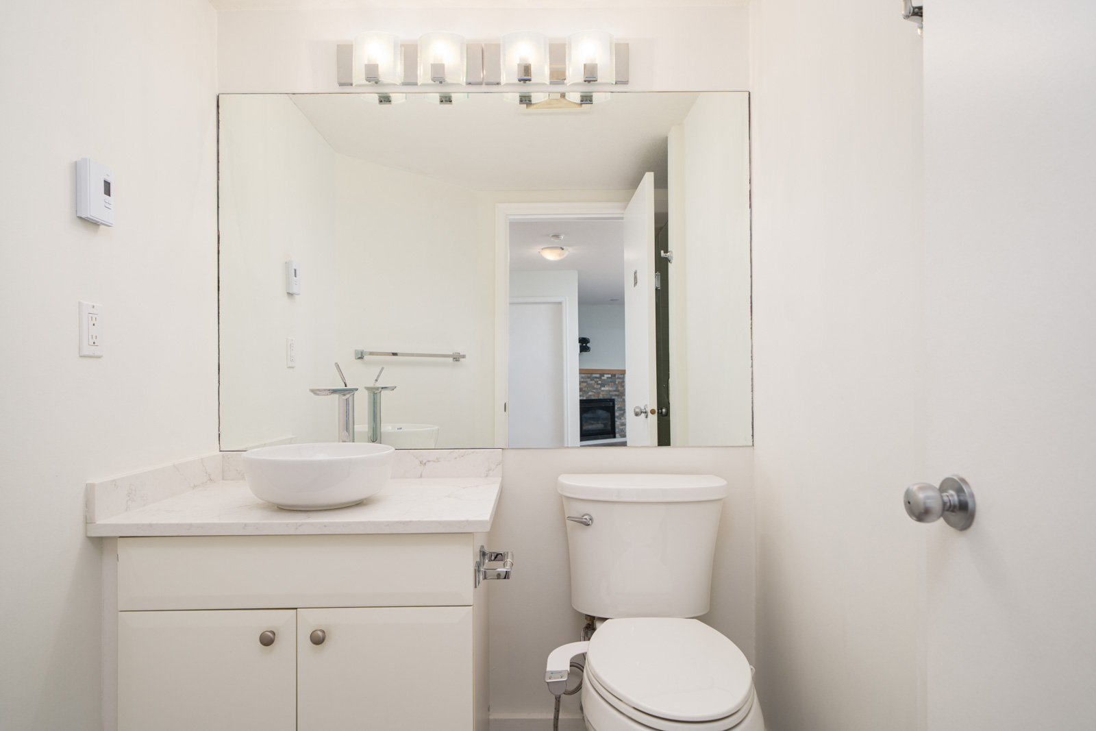 A small white bathroom features a vanity with a vessel sink, a toilet, a large mirror, and overhead lights. The door is open, showing a glimpse of another room.