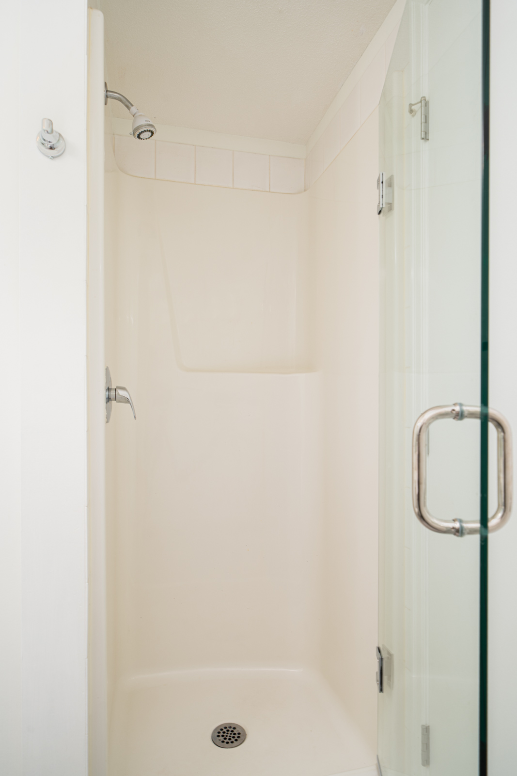 A small, clean, beige shower stall with a glass door, wall-mounted showerhead, and a built-in shelf.