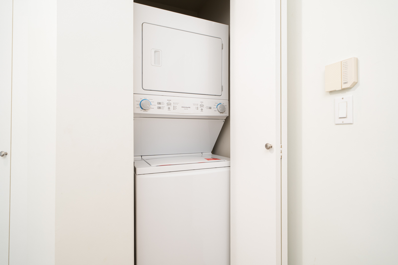Stacked washer and dryer unit in a small laundry closet with white folding doors, adjacent to a light switch and thermostat on the wall.
