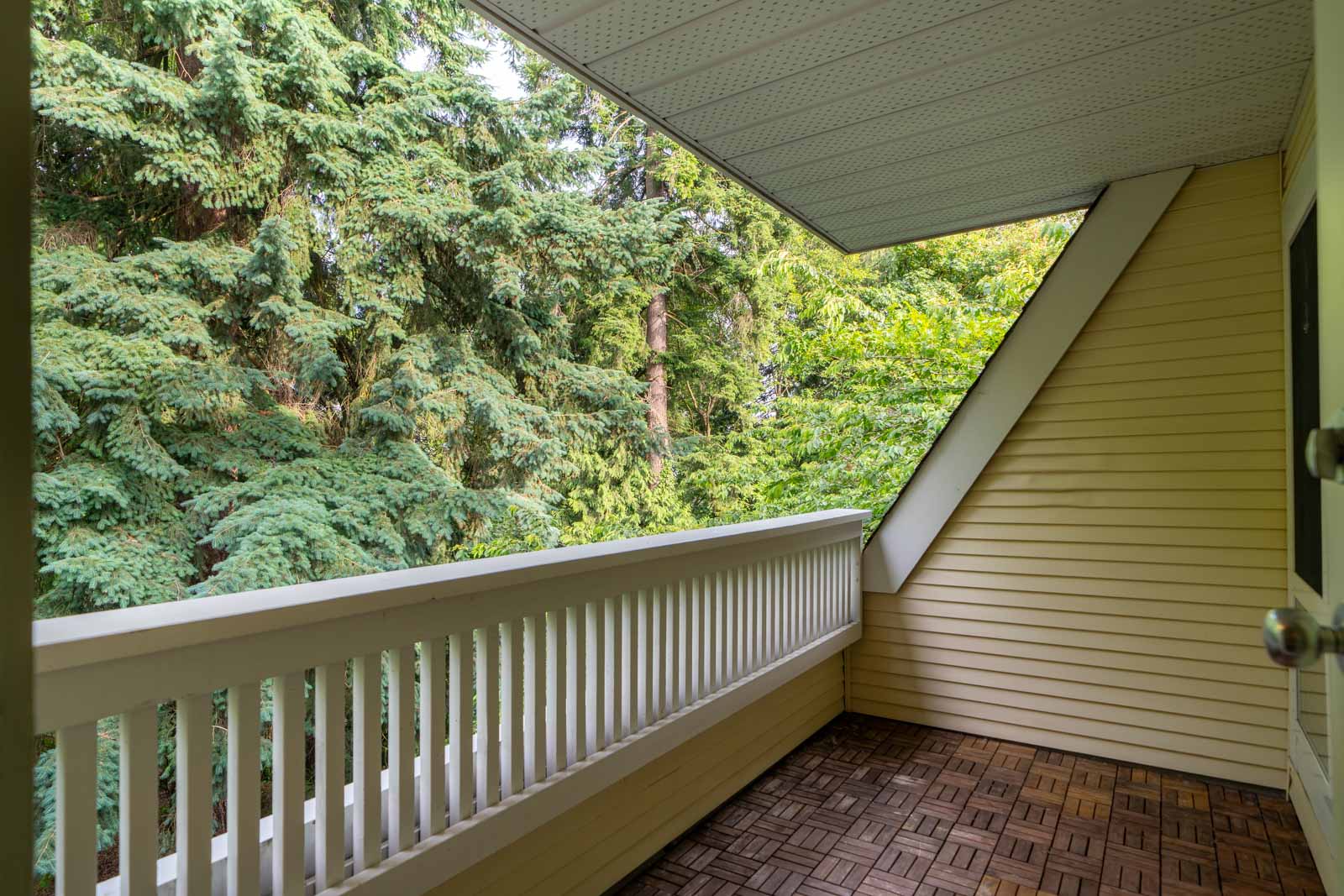 A covered balcony with a wooden floor and railing overlooks a dense area of tall green trees.