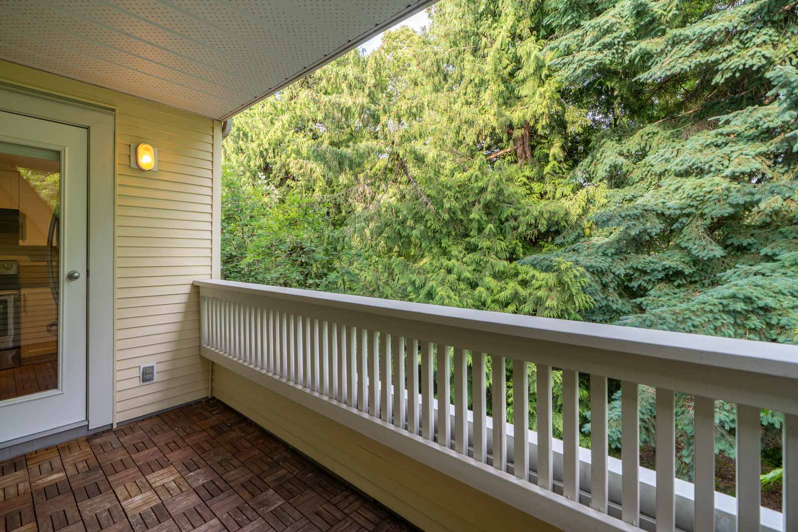 A covered balcony with wooden flooring, white railing, and a view of dense green trees. A door and light are visible on the left side.