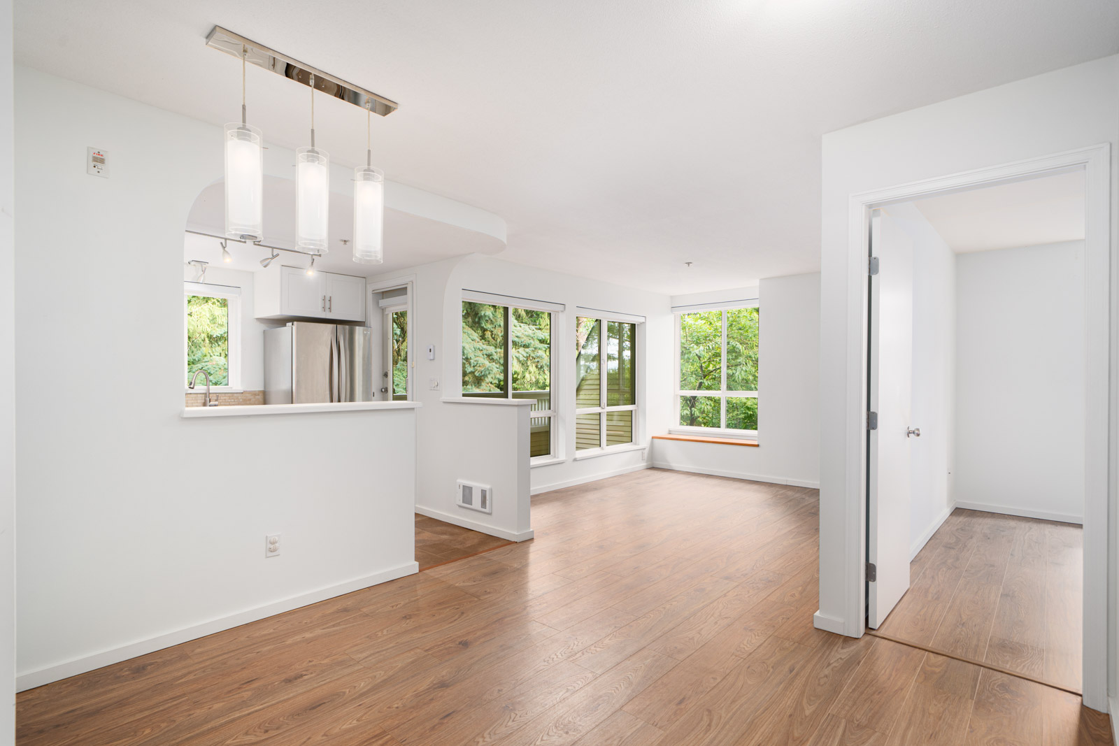 Open-plan apartment interior with wood flooring, white walls, modern light fixture, kitchen with stainless steel appliances, large windows, and an adjacent room with a closed door.