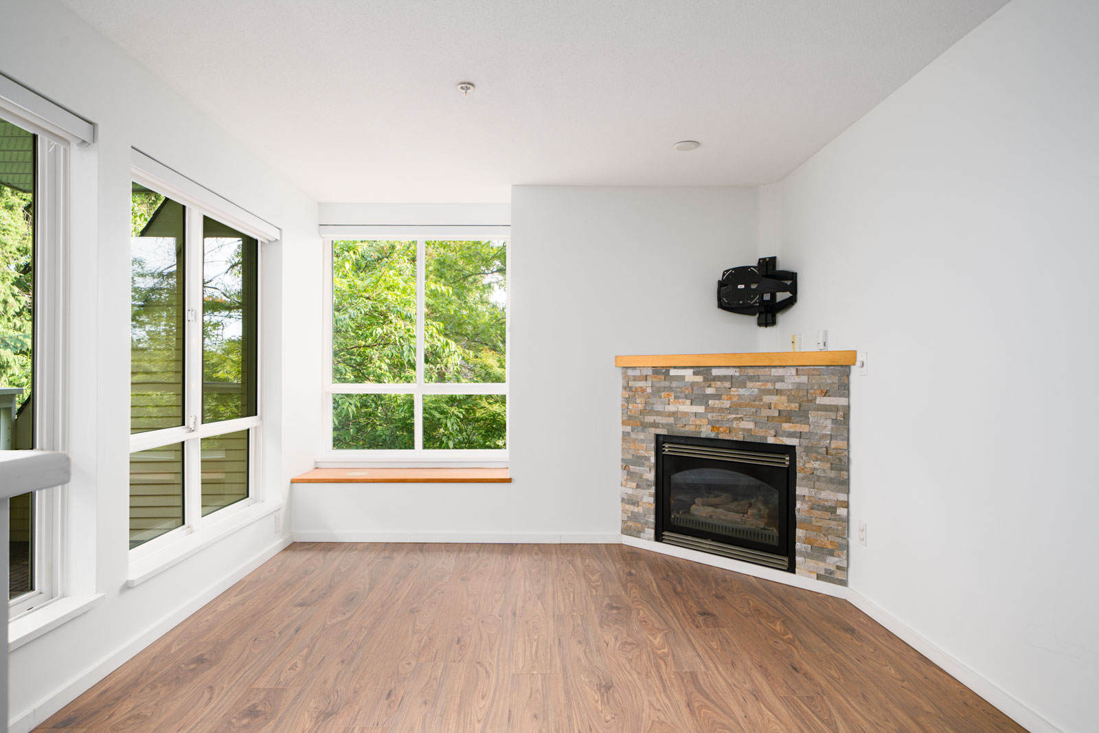 Empty living room with large windows, wood-style flooring, a stone-faced fireplace in the corner, and a wall mount for a TV above the fireplace.