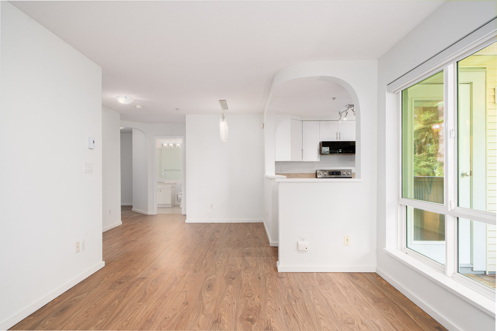 Bright, unfurnished apartment interior with white walls, wood flooring, open kitchen, and large windows overlooking greenery. Bathroom visible through doorway in the background.