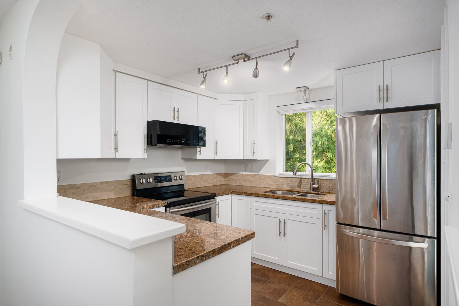 Modern kitchen with white cabinets, stainless steel appliances, granite countertops, track lighting, and a window above the sink with a view of greenery.