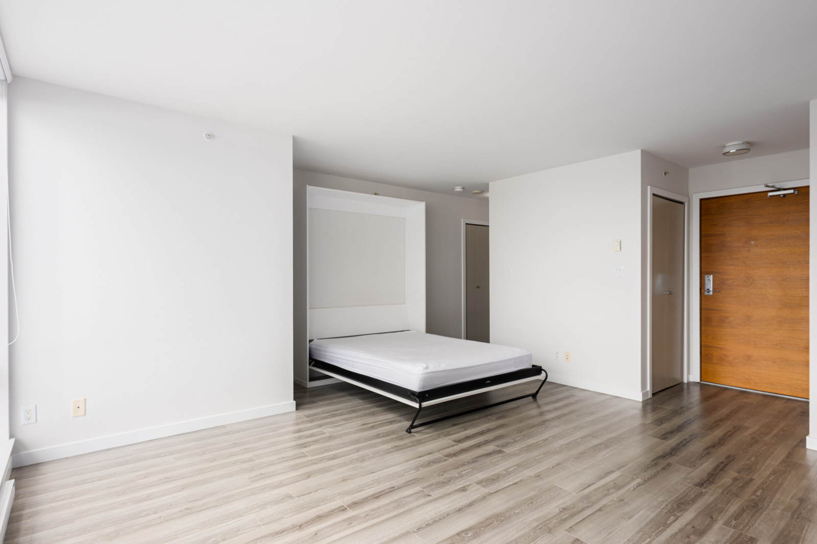 A minimalist room with light wood flooring, a fold-out bed, white walls, and a wooden door. The space appears uncluttered and well-lit by natural light.