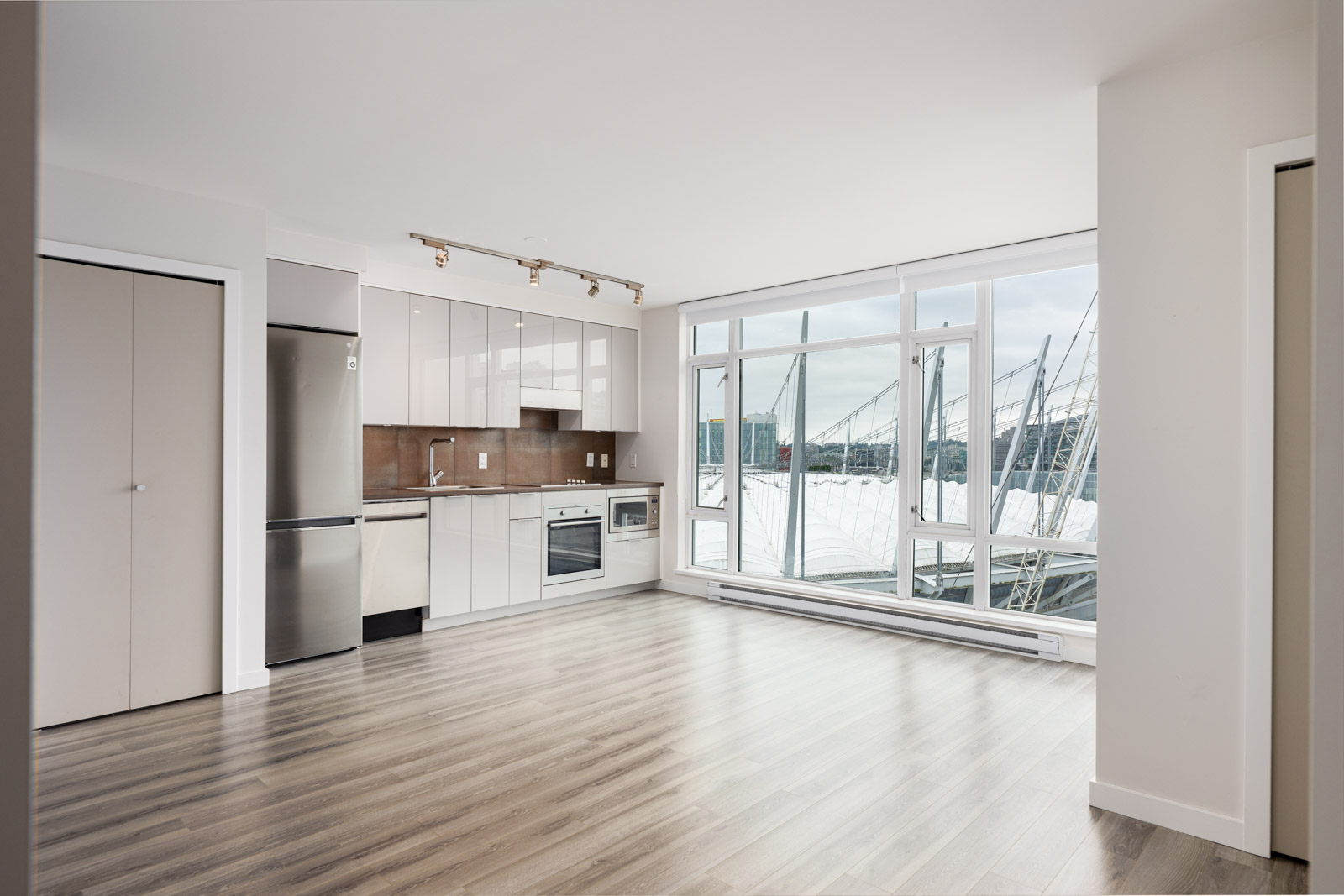 Modern, unfurnished apartment with wood flooring, stainless steel appliances, and large windows overlooking boats and water outside.