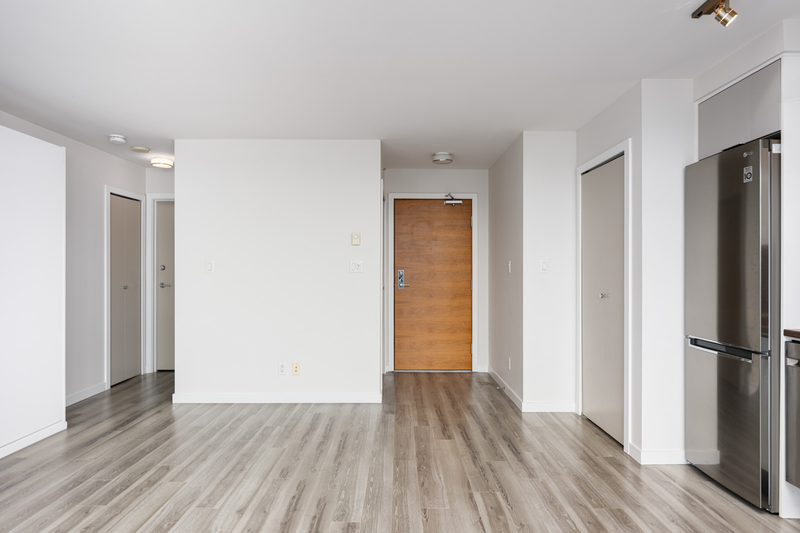 Interior of an empty modern apartment with light-colored walls, wood flooring, a wooden entrance door, and a stainless steel refrigerator on the right.