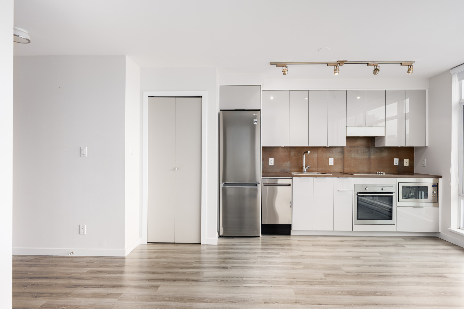 Modern kitchen with white cabinets, stainless steel appliances, light wood flooring, brown backsplash, and a closed closet door. Room is empty and well-lit by natural light.