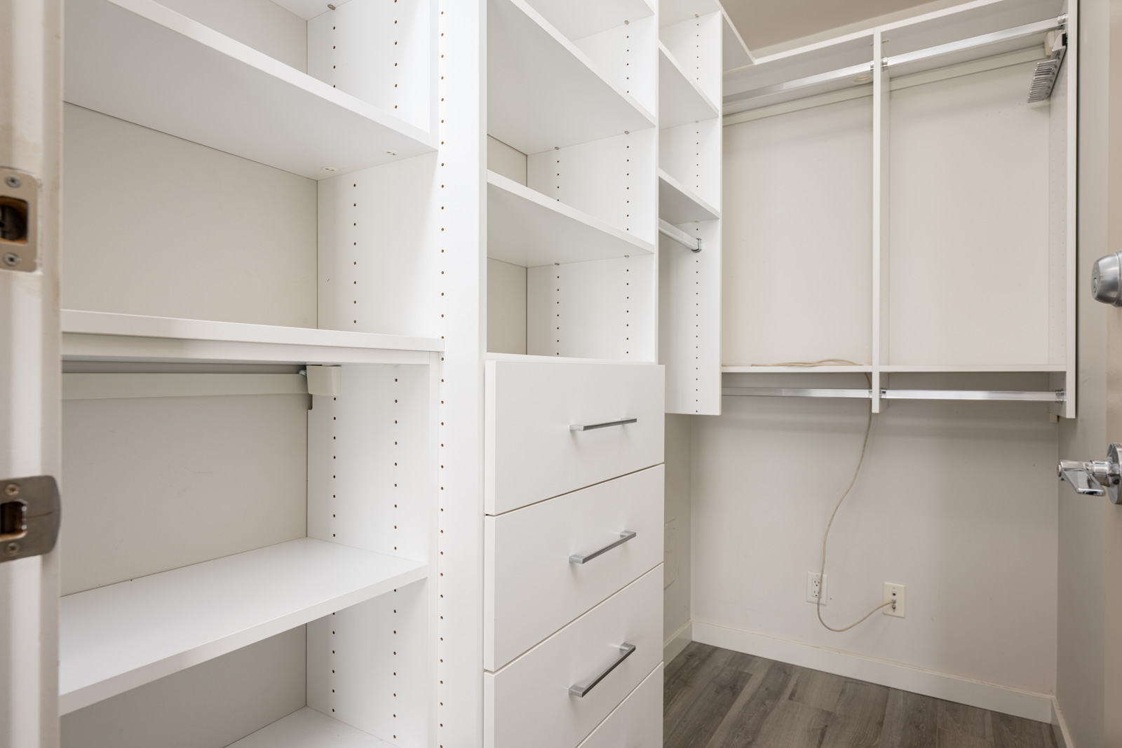 Walk-in closet with white built-in shelves, drawers, and hanging rods, featuring light-colored walls and a gray wood floor. An electrical outlet is visible near the baseboard.