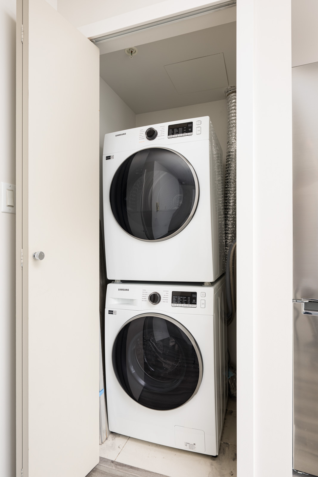 Stacked Samsung washer and dryer units are installed in a small laundry closet with a partially open door.